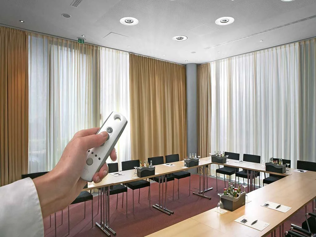 A hand holding a remote control, opening and closing a set of motorized curtains in a modern conference room